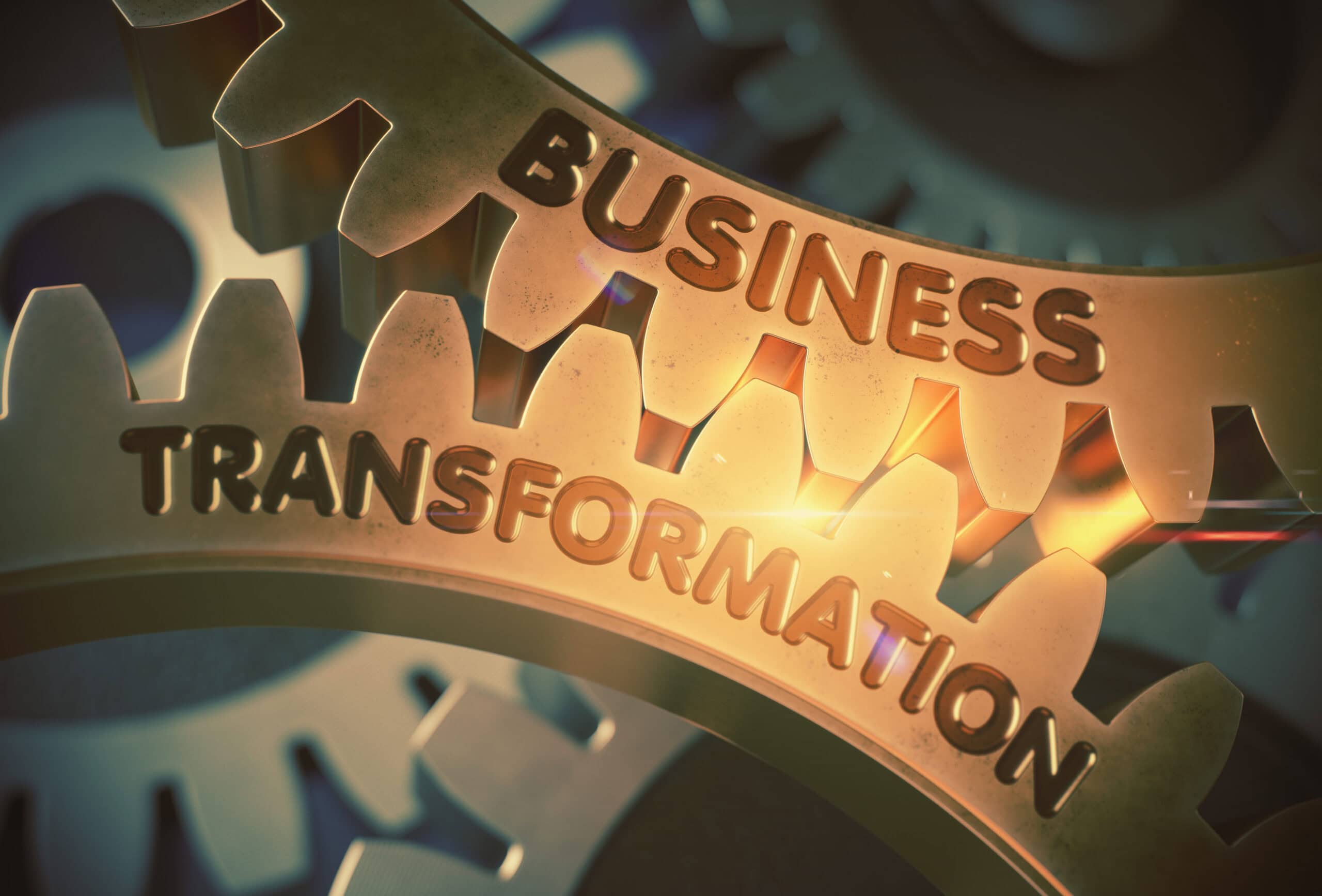 Auf dem Bild sind gro&szlig;e metallische Zahnr&auml;der zu sehen, die ineinandergreifen. Auf dem vorderen Zahnrad steht das Wort &sbquo;Transformation&lsquo;, auf dem hinteren das Wort &sbquo;Business&lsquo;. Warme Lichtreflexe betonen die goldene Oberfl&auml;che der Zahnr&auml;der.