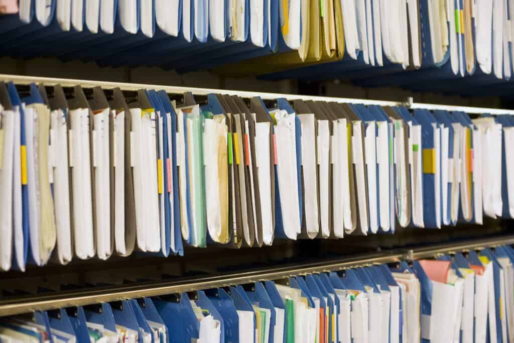 n mehreren Regalen h&auml;ngen zahlreiche Ordner und Mappen nebeneinander. Die Unterlagen sind eng gesteckt, teilweise farblich sortiert. Die Szene wirkt wie ein Archiv oder ein B&uuml;ro mit vielen Papierakten.