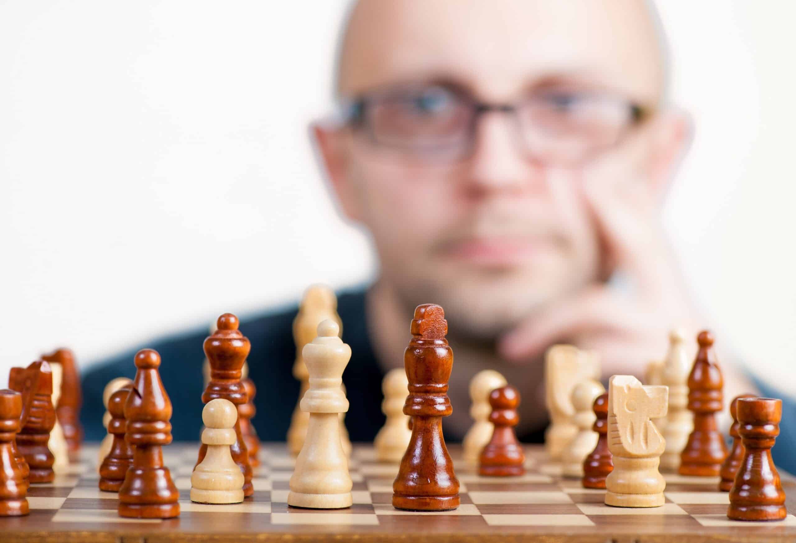 Im Vordergrund steht ein Schachbrett mit hellen und dunklen Figuren in Spielaufstellung. Im Hintergrund ist eine Person unscharf zu sehen, die mit der Hand das Gesicht st&uuml;tzt und auf das Schachbrett schaut.