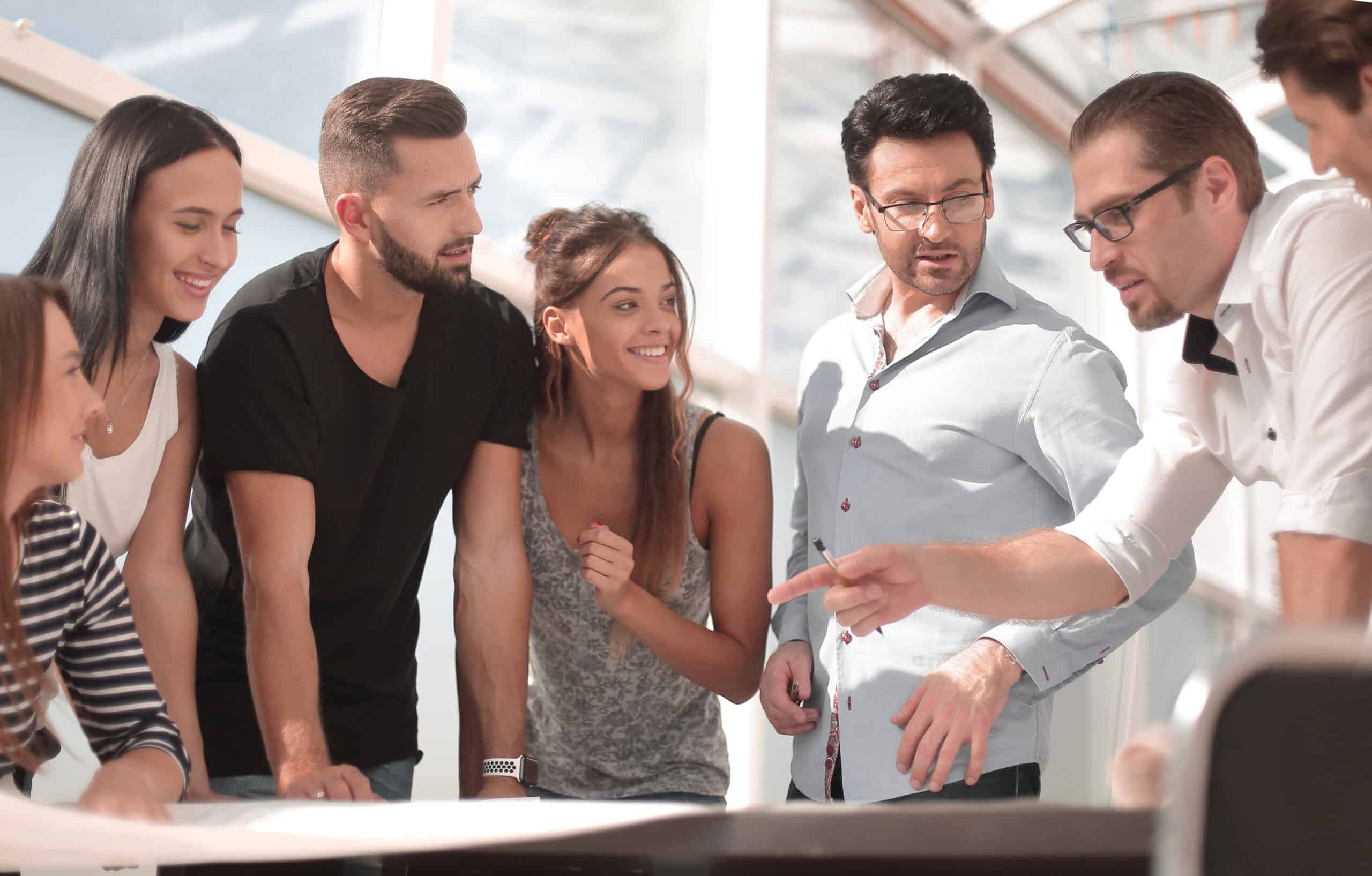 Eine Gruppe aus sechs Personen steht dicht beisammen um einen Tisch in einem hellen Raum. Ein Mann mit Brille zeigt auf etwas auf dem Tisch, w&auml;hrend die anderen nach vorne blicken oder miteinander sprechen. Die Stimmung wirkt konzentriert und kooperativ