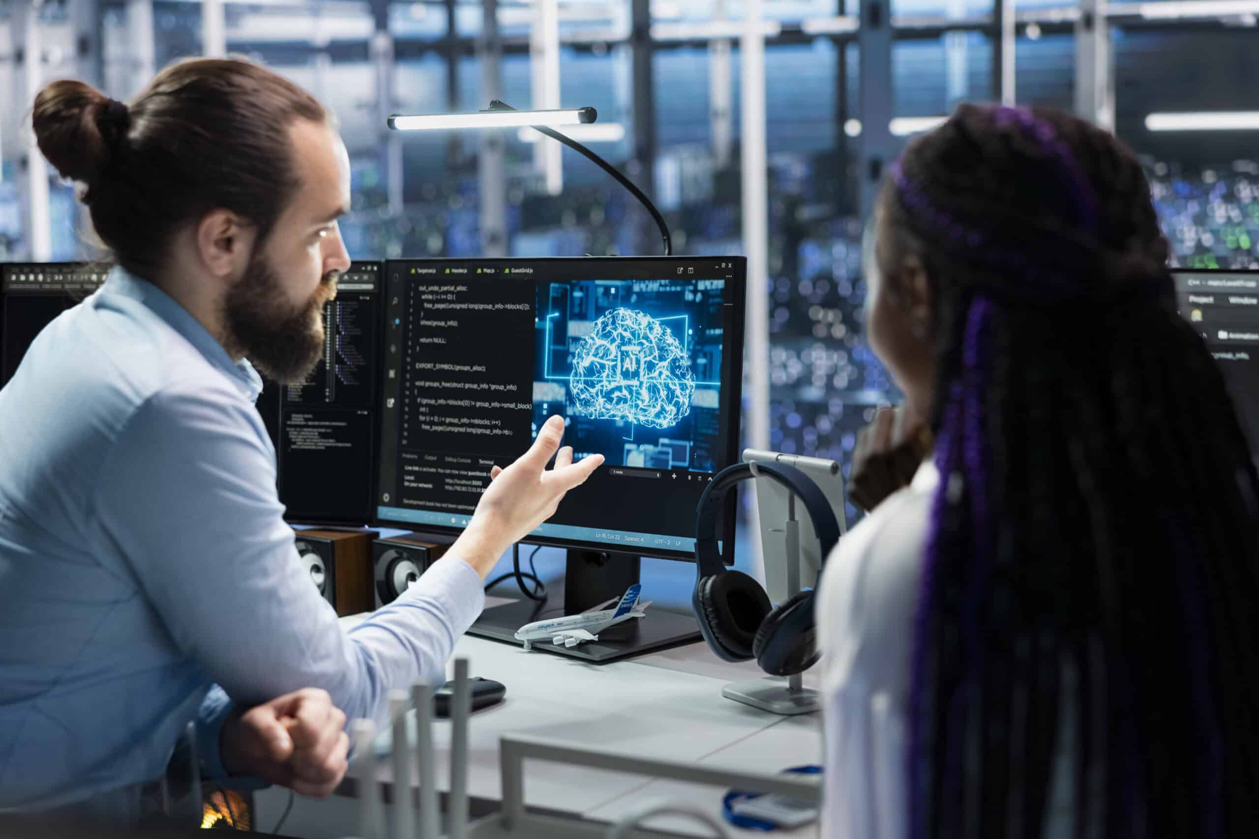 Zwei Personen sitzen vor Monitoren mit technischem Code und einer grafischen Darstellung; eine Person zeigt auf den Bildschirm.