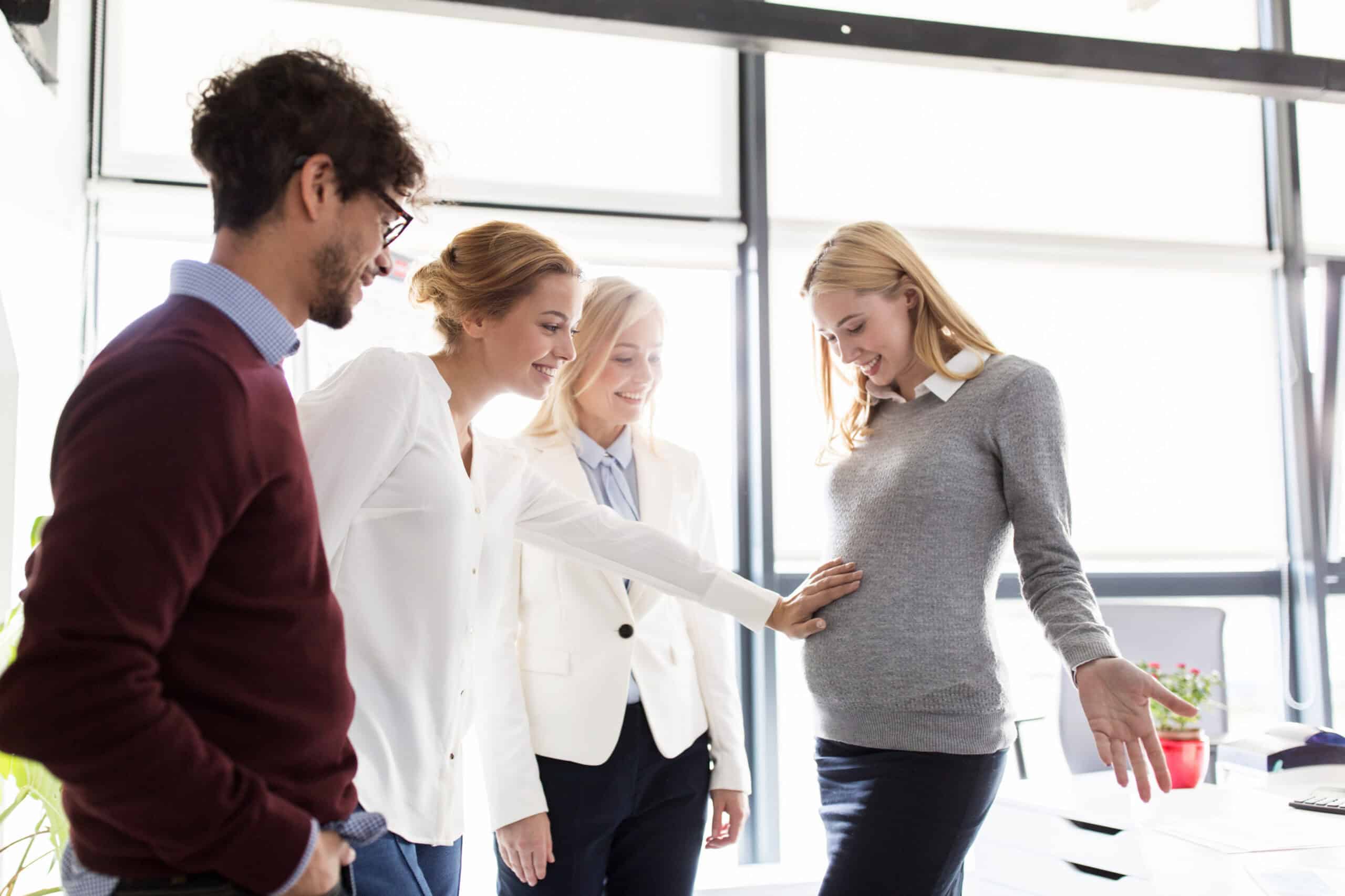 Vier Personen stehen in einem hellen B&uuml;ro gemeinsam beisammen. Eine Person legt behutsam eine Hand auf den Bauch einer anderen Person, die offensichtlich schwanger ist. Alle l&auml;cheln oder schauen freundlich. Im Hintergrund sind gro&szlig;e Fenster und B&uuml;rom&ouml;bel zu sehen.
