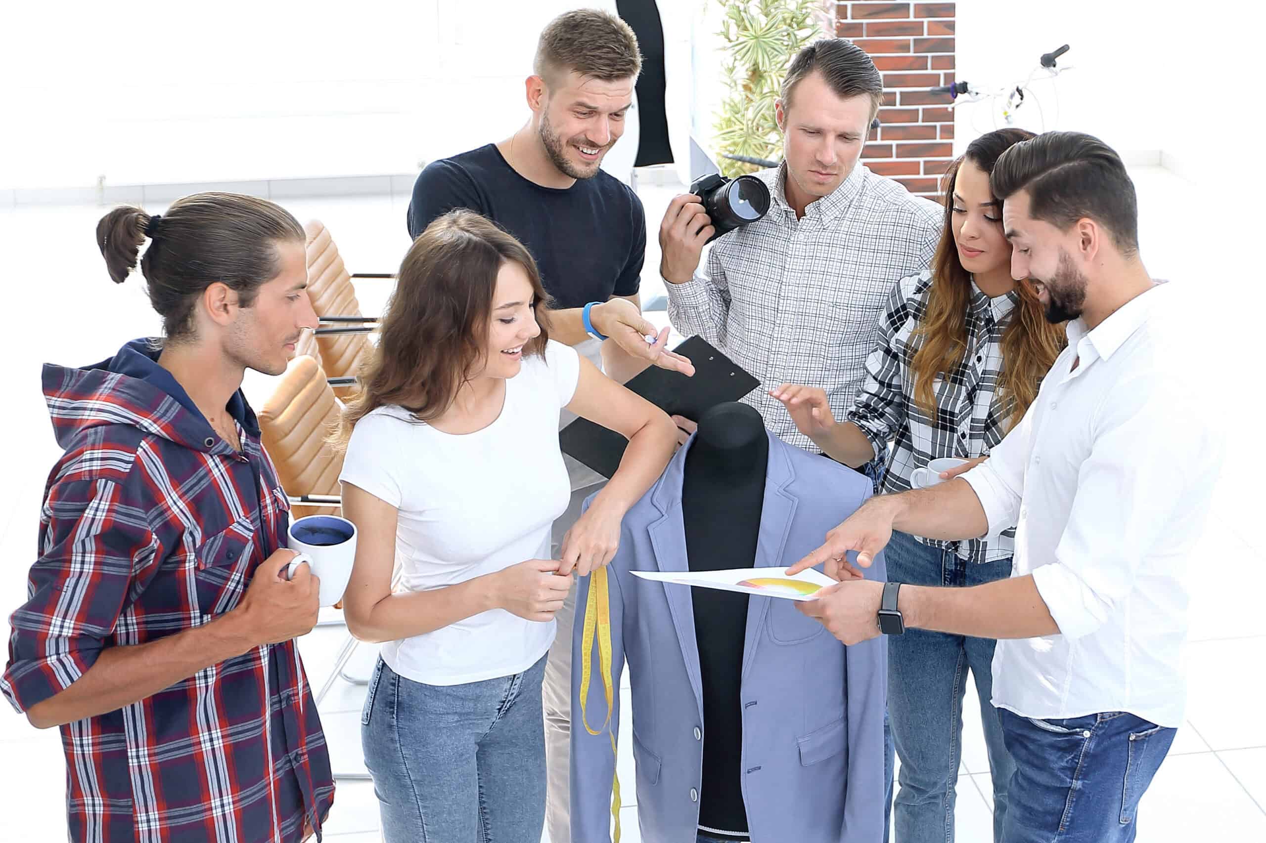 Sechs Personen stehen in einem hellen Raum um eine Schneiderpuppe, die mit einem Jackett bekleidet ist. Eine Person h&auml;lt ein Blatt Papier mit Farbdarstellungen hoch, w&auml;hrend die anderen darauf zeigen oder interessiert darauf blicken. Eine Person h&auml;lt eine Kamera, eine andere eine Tasse. Die Stimmung wirkt kreativ und kooperativ.