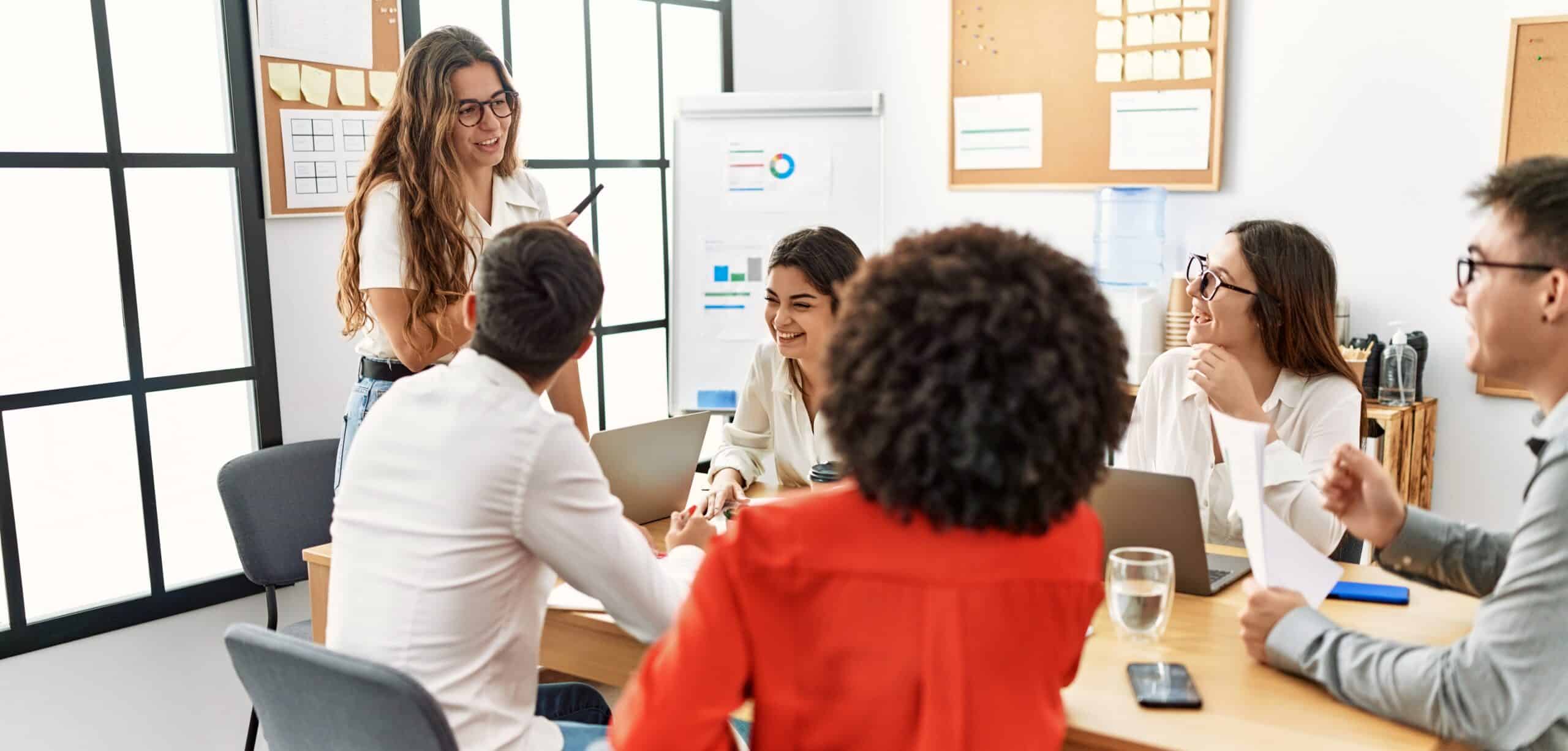 Eine Gruppe von Menschen sitzt in einem hellen Besprechungsraum an einem Tisch. Eine Person steht vorne, l&auml;chelt und spricht zur Gruppe. Die anderen Personen sitzen mit Laptops, Notizen und Getr&auml;nken am Tisch und h&ouml;ren zu. Im Hintergrund sind gro&szlig;e Fenster, Pinnw&auml;nde und ein Flipchart.
