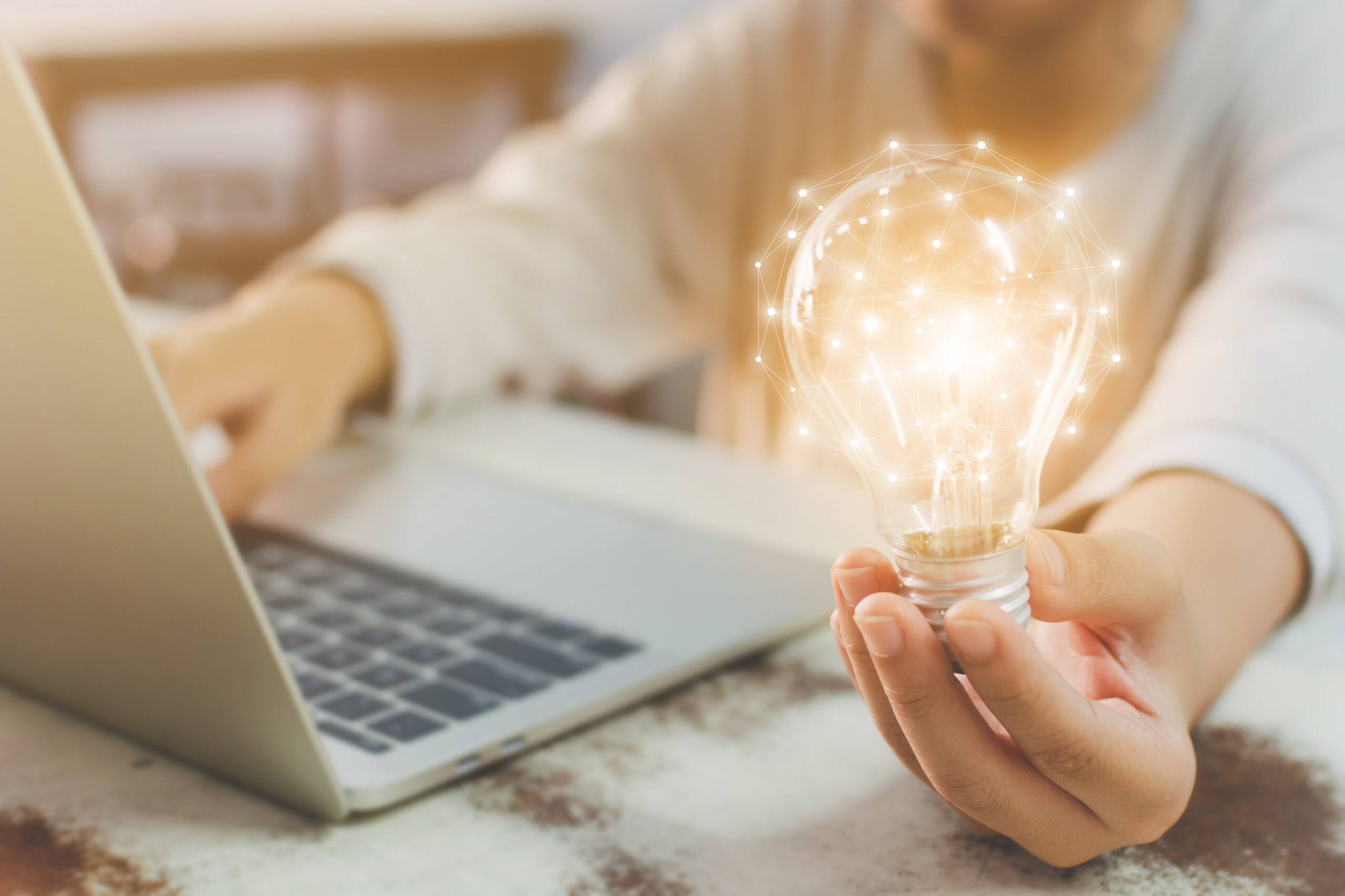 Eine Person h&auml;lt eine hell leuchtende Gl&uuml;hbirne, deren Oberfl&auml;che durch feine Lichtpunkte und Linien wie ein digitales Netzwerk wirkt. Im Hintergrund steht ein aufgeklappter Laptop. Das Bild vermittelt Innovation, Qualit&auml;t und die Verbindung aus moderner Technologie und durchdachter Arbeitsweise.