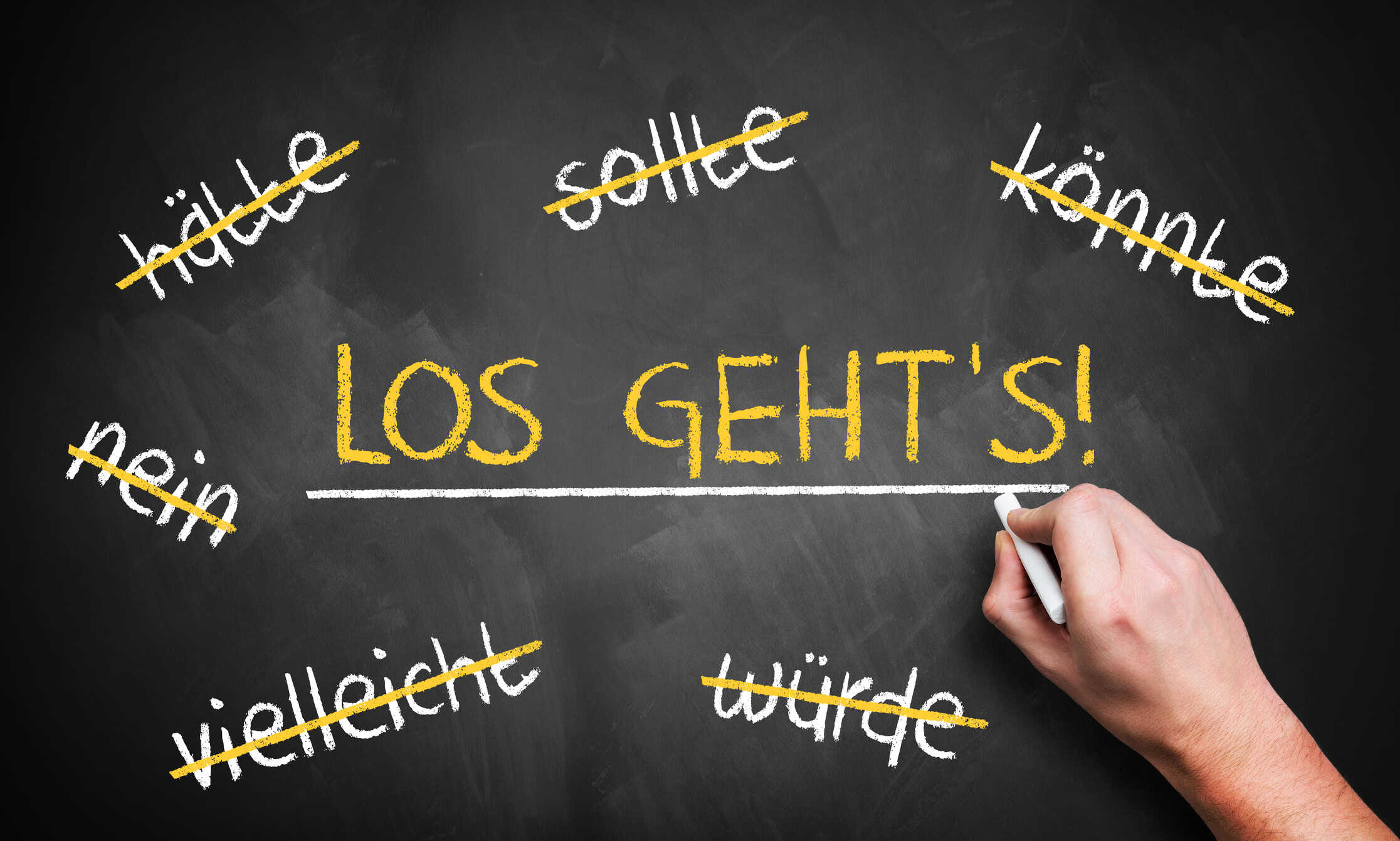 Auf einer Tafel steht gro&szlig; &sbquo;Los geht&rsquo;s!&lsquo;; darum herum sind W&ouml;rter wie &sbquo;h&auml;tte&lsquo;, &sbquo;sollte&lsquo; und &sbquo;vielleicht&lsquo; durchgestrichen. Eine Hand schreibt mit Kreide.