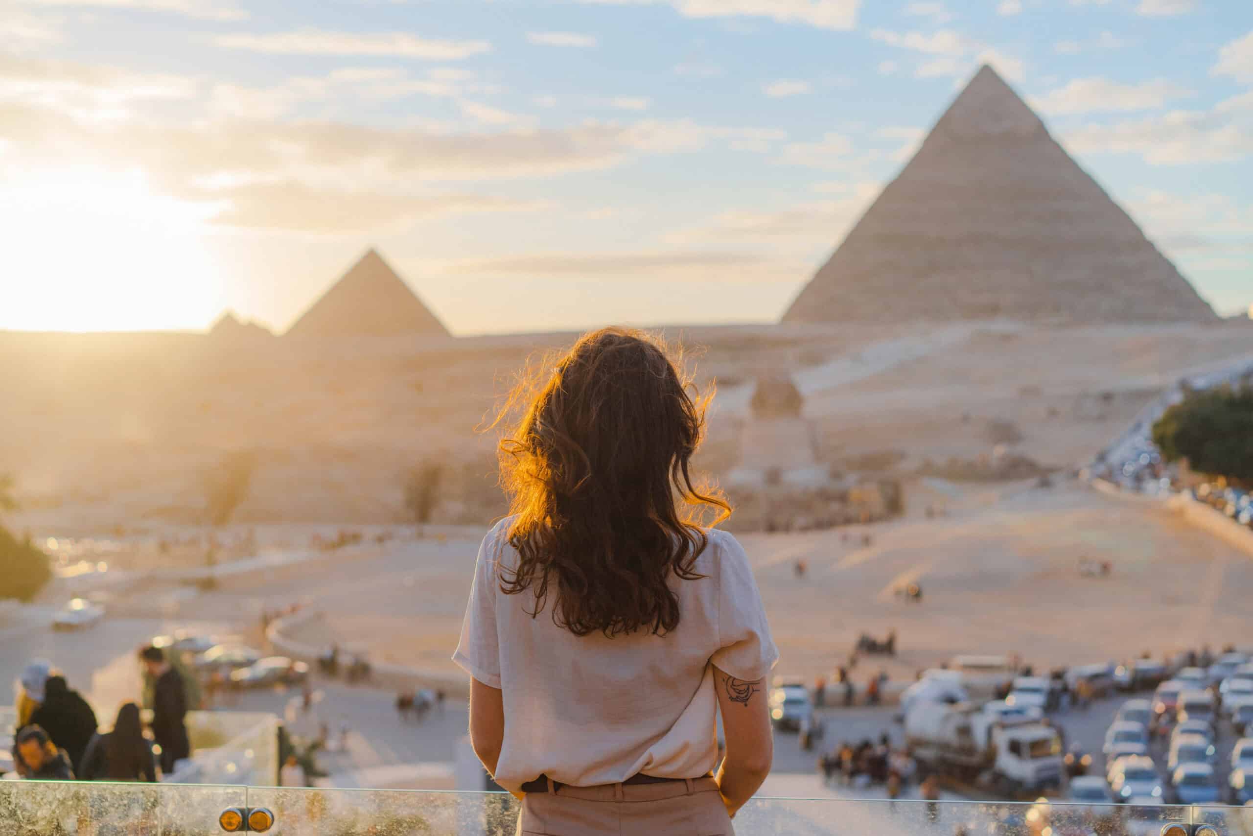 Eine Person steht mit dem R&uuml;cken zur Kamera auf einer erh&ouml;hten Plattform und schaut auf die Pyramiden in einer W&uuml;stenlandschaft. Die Sonne steht tief und beleuchtet die Szene warm. Im Vordergrund bewegen sich Menschen und Fahrzeuge.