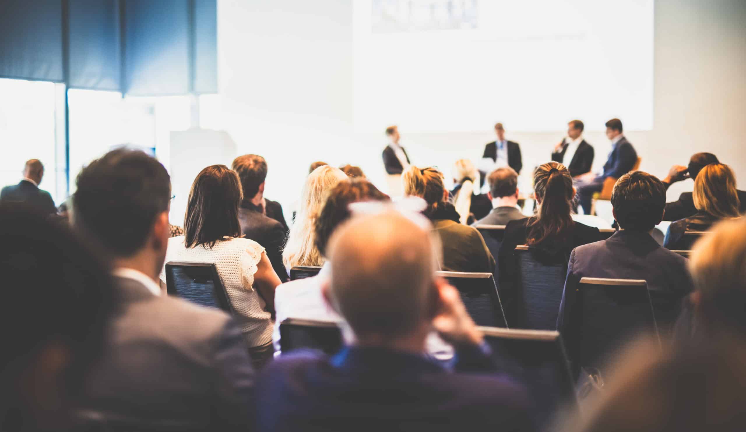 In einem hellen Konferenzraum sitzt ein gr&ouml;&szlig;eres Publikum und blickt nach vorn auf mehrere Personen, die auf einer erh&ouml;hten B&uuml;hne oder Panel-Fl&auml;che sitzen und sprechen. Die Zuh&ouml;rer sind von hinten zu sehen, w&auml;hrend die B&uuml;hne und die Vortragenden leicht unscharf im Hintergrund erscheinen.