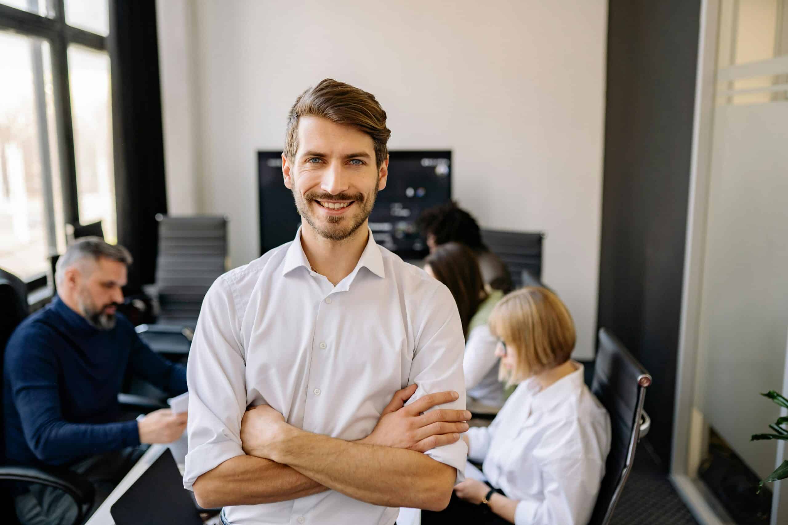 Im Vordergrund steht eine Person mit verschr&auml;nkten Armen und l&auml;chelt in die Kamera. Im Hintergrund sitzen drei weitere Personen an einem Tisch und arbeiten gemeinsam an Unterlagen oder einem Laptop. Der Raum wirkt modern und hell.