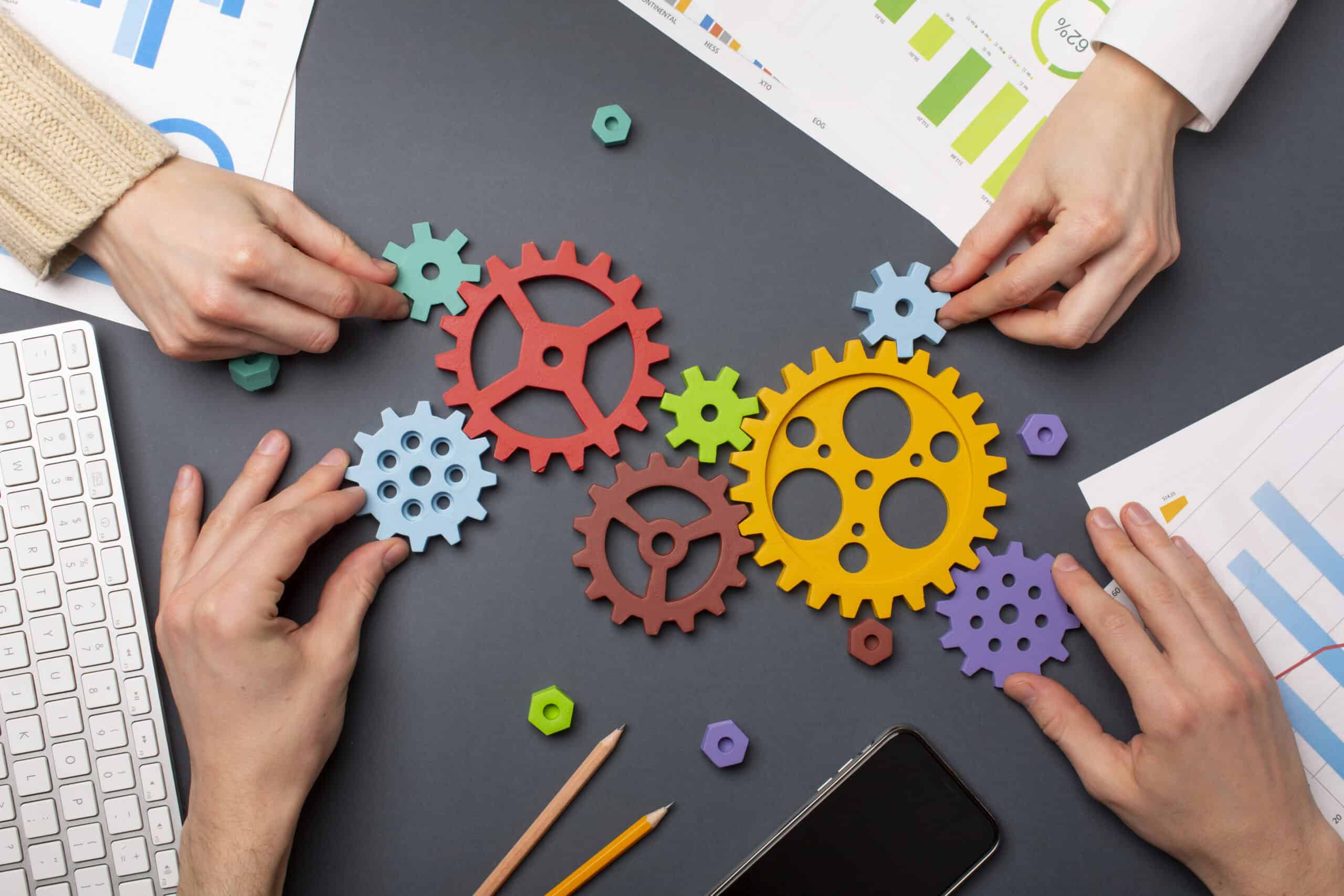 Das Bild zeigt mehrere H&auml;nde, die farbige Zahnr&auml;der gemeinsam so arrangieren, dass sie zu einem funktionierenden System zusammenfinden. Auf dem Tisch liegen Diagramme, Auswertungen und digitale Arbeitsmittel &ndash; ein klarer Hinweis auf strukturierte Zusammenarbeit und gemeinsames Gestalten von Prozessen. Die Szene vermittelt koordiniertes Teamwork, Verstehen von Zusammenh&auml;ngen und das bewusste Verzahnen einzelner Elemente zu einem starken Ganzen.