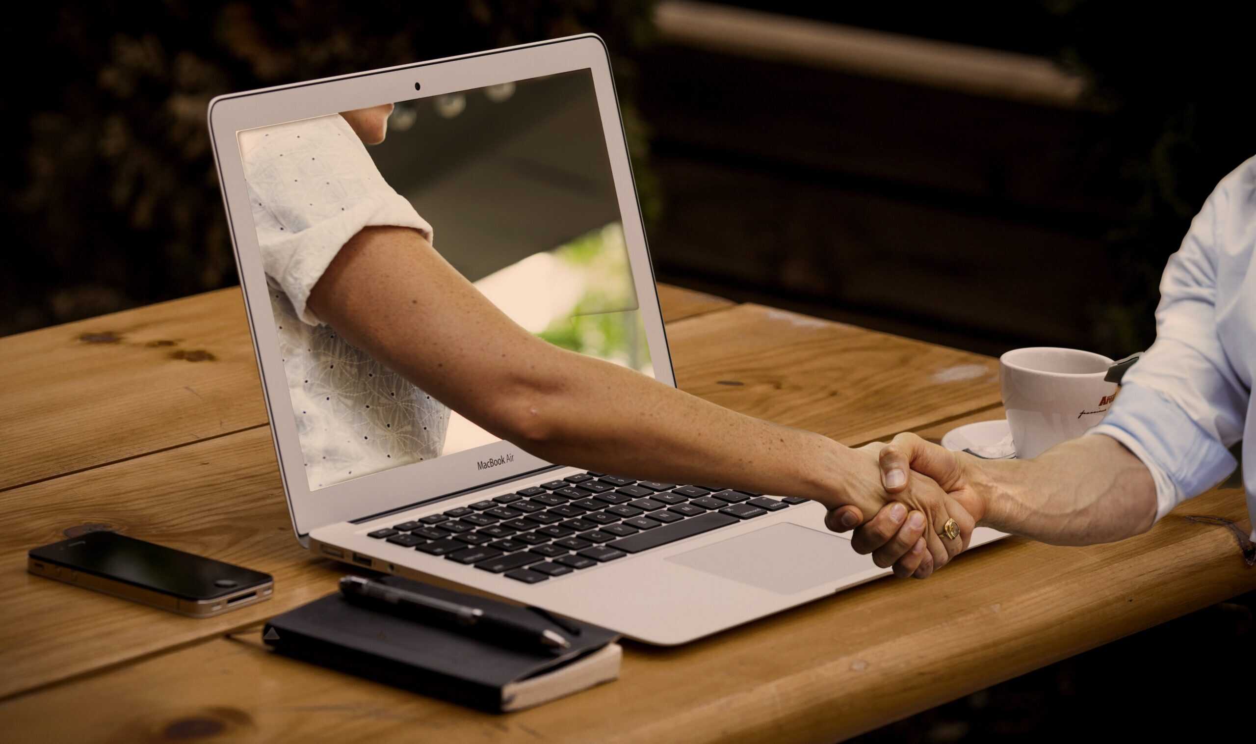 Zwei Personen geben sich die Hand; ein Arm erscheint aus dem Bildschirm eines Laptops.